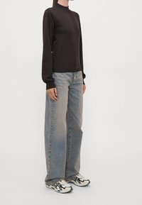 Person in einem schwarzen Langarmoberteil, hellblauen weiten Jeans und weißen und schwarzen Sneakers, der vor einem schlichten Hintergrund steht.