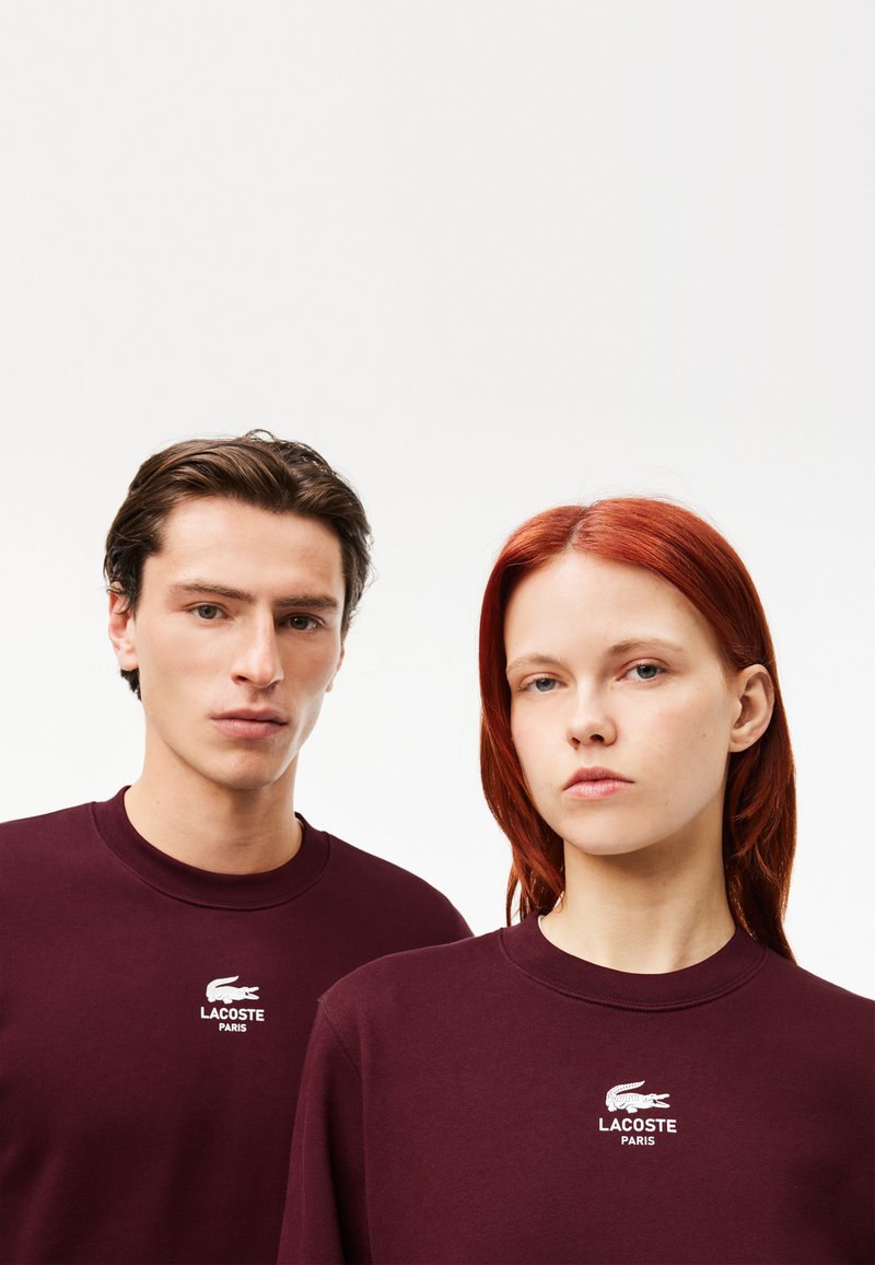 Sweatshirt bordeaux arborant le logo Lacoste brodé. Conception à encolure ronde avec une texture douce et une coupe décontractée, porté par deux modèles.