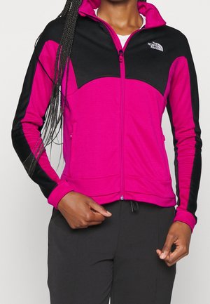 Woman wearing a magenta and black zip-up North Face jacket with long sleeves and black pants, standing with hands near waist.
