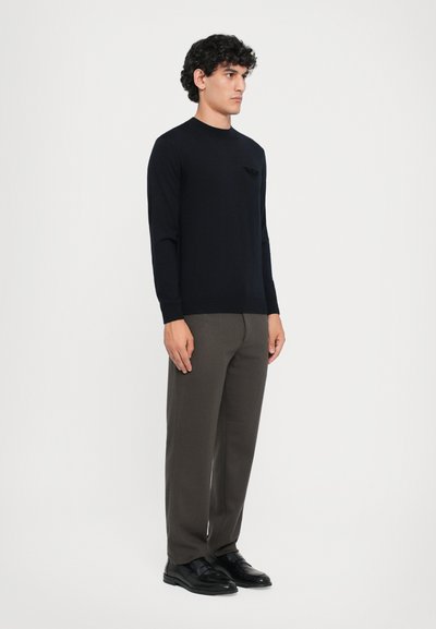 Pulover tricotat bleumarin cu gât rotund și detaliu texturat cu logo, asociat cu pantaloni gri și pantofi negri, are mâneci lungi și un design minimalist.