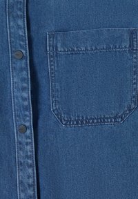 Gros plan sur un tissu denim bleu montrant une poche poitrine avec des coutures visibles et deux boutons-pression noirs sur la patte de boutonnage d'une chemise.