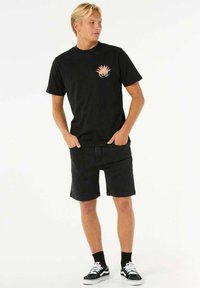 Zwarte katoenen t-shirt met een kleurrijke grafische print op de borst, gecombineerd met zwarte denim shorts en zwarte instappers met witte accenten.