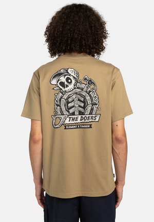 Camiseta de manga corta color beige con un diseño gráfico en negro en la parte posterior, que representa una calavera con herramientas y el texto "¡EL ELEMENTO DE LOS HACEDORES X TIMBER!"