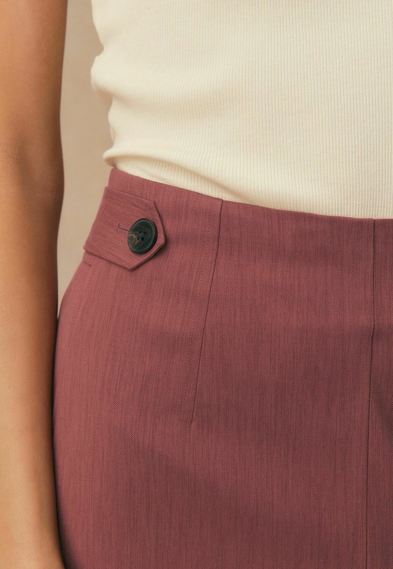 Rok in hoge taille in mauve met een knoopdetail aan de zijkant, gecombineerd met een crème ribgebreide mouwloze top die in de rok is gestopt, gedragen door het bovenlichaam van een persoon.