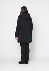 Giacca invernale nera con cappuccio, caratterizzata da un taglio leggermente più lungo, una texture elegante e una tasca con zip laterale sulla manica.
