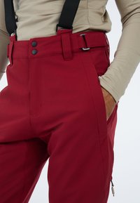 Rode outdoorbroek met een gladde textuur, voorzien van knoop- en riemslot-sluitingen, zijzakken en verstelbare zwarte bretels.
