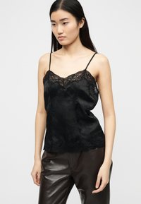 Νεαρή γυναίκα με μακριά σκούρα μαλλιά, φορώντας μαύρο τοπ με δαντέλα και σκουρόχρωμο δερμάτινο παντελόνι, κοιτάζοντας ελαφρώς προς τα πλάγια.