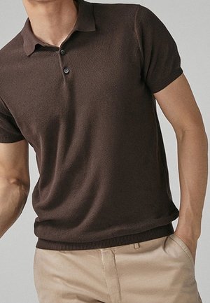 Mężczyzna w ciemnobrązowej, krótkim rękawem, dzianinowej koszulce polo włożonej do beżowych spodni na neutralnym tle.