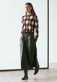 Pullover mit einem Rautenmuster in Kastanienbraun, Creme und Schwarz; kombiniert mit einem langen schwarzen Lederrrock und kniehohen schwarzen Stiefeln.