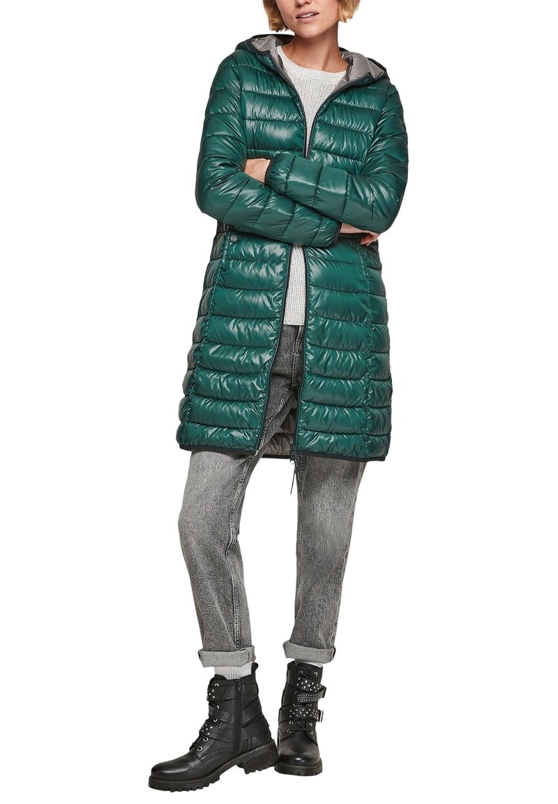 Grøn quilted puffer jakke med hætte, der har en frontal lynlås. Båret over en lys sweater og grå opsmøgede jeans, parret med sorte støvler.