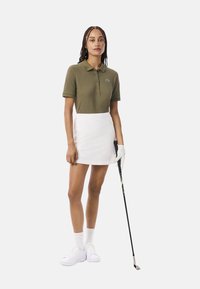 Polo de color verde oliva con mangas cortas, falda blanca, zapatos blancos y un palo de golf. Tejido texturizado, sutil acento de logotipo en la camiseta.