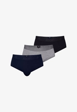 Homme debout pieds nus devant un fond uni, portant un slip noir avec un logo sur la ceinture, face à l'avant avec une posture détendue.