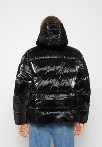Černá péřová bunda s lesklým povrchem, vybavená kapucí, prošívaným designem a oversized siluetou. Texturovaná tkanina s viditelnými švy.