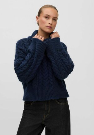 Femme portant un pull en maille épaisse bleu marine avec des motifs torsadés, tenant le col, associé à un pantalon noir sur fond uni.