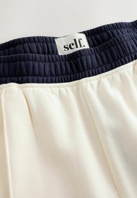 Baltos sportinės kelnės su tamsiai mėlynu elastingu juosmeniu, ant kurio yra etikėtė su užrašu „self.“ Audinys atrodo minkštas ir šiek tiek tekstūruotas.