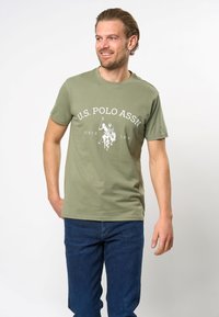 Olīvu zaļas krāsas kokvilnas t-krekls ar baltu U.S. Polo Assn. logotipu un grafisko zīmējumu. īsas piedurknes un apaļš kakla izgriezums. Vilkts kopā ar tumši ziliem džinsiem.
