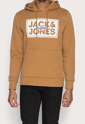 Mand iført en brun hættetrøje med teksten "JACK & JONES Original" på fronten og sorte bukser.