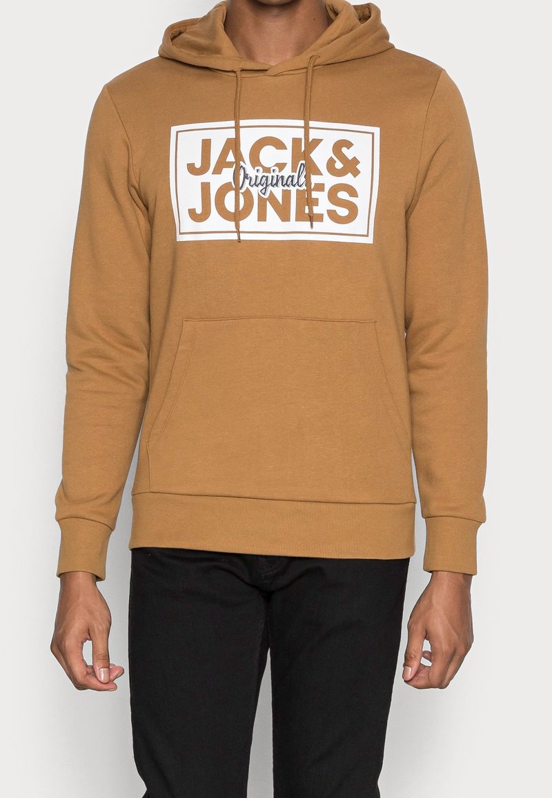 Homme portant un sweat à capuche marron avec le texte « JACK & JONES Original » sur le devant et un pantalon noir.