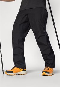 Črne lahke hlače s slim fit krojem in kontrastnim robom na strani, v kombinaciji z oranžno-sivimi čevlji za tek po poteh, ki imajo teksturirane podplate.