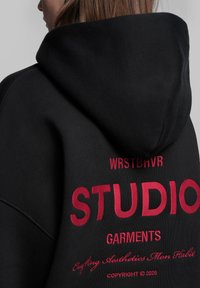 Felpa con cappuccio nera caratterizzata da testo ricamato rosso: "WRSTBVR STUDIO GARMENTS" e ulteriore slogan, con una morbida texture del tessuto.