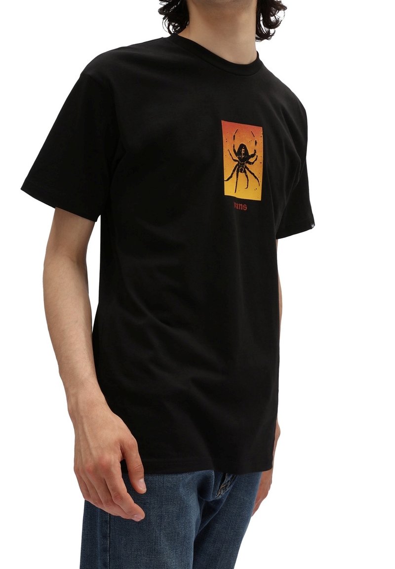 Personne portant un T-shirt noir avec un carré dégradé du rouge au jaune et un graphique d'araignée noire au centre, associé à un jean bleu.