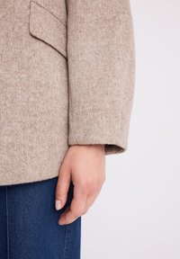 Beżowa marynarka z mieszanki wełny o teksturowanym wykończeniu, z kieszenią naszytą na rękawie, w połączeniu z ciemnoniebieskimi dżinsami.