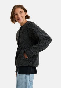 Grijze, gestructureerde fleece jas met een relaxte pasvorm, twee zijzakken en een ronde kraag, gelaagd over een zwart shirt en lichte spijkerbroek.