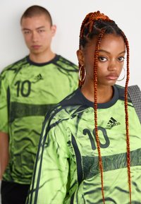 To personer iført matchende lysegrønne Adidas-drakter med nummer 10, en med fletninger og hoop øredobber i forgrunnen, en uskarp i bakgrunnen.