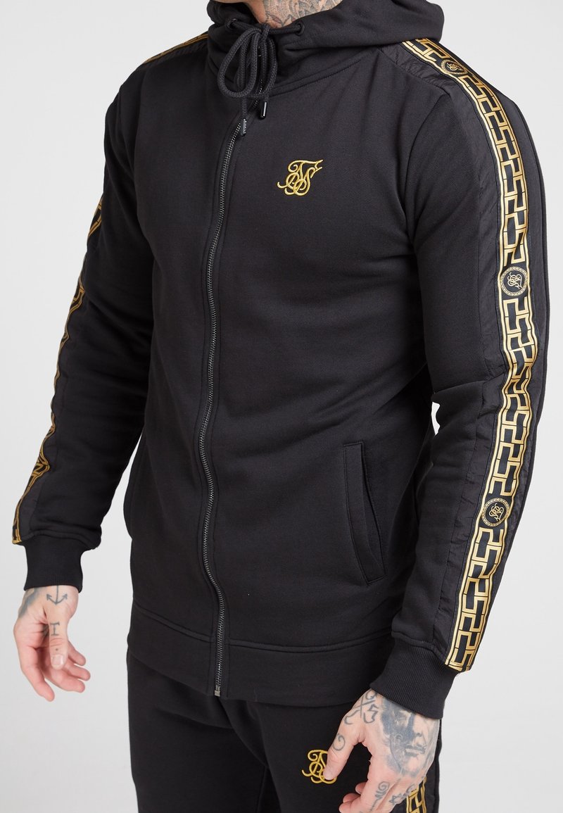 Felpa nera con zip e dettagli dorati, logo, strisce laterali con motivo, cappuccio con coulisse e tasche anteriori. Realizzata in tessuto morbido.