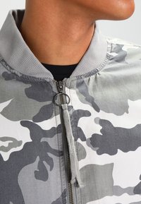 Chaqueta bomber de camuflaje gris con cuello de canalé, cremallera frontal y detalle de cordón. Presenta una textura de tela suave y un diseño moderno.