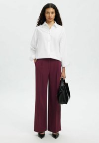 Femme debout aux cheveux bouclés, portant une chemise blanche boutonnée, un pantalon large bordeaux, des chaussures noires à bouts pointus, tenant un sac à main noir.