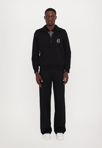Felpa nera con zip fino a un quarto e logo, abbinata a pantaloni neri a gamba larga e sneakers bianche, su uno sfondo neutro.