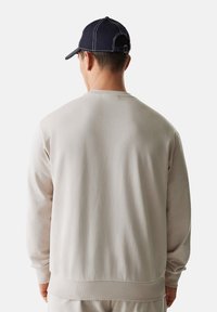 Felpa grigia chiara con collo rotondo e polsini a costine. Un cappellino blu navy con cuciture bianche completa l'outfit. Texture complessivamente liscia.