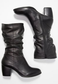 Botas de cuero negro hasta el tobillo con diseño arrugado, punta redonda y tacón apilado. Presentan una cremallera lateral para facilitar el calzado. Textura suave.