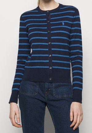 Vrouw draagt een marineblauw en blauw horizontaal gestreepte cardigan met knopen, gecombineerd met donkerblauwe jeans, staand met ontspannen armen langs het lichaam.