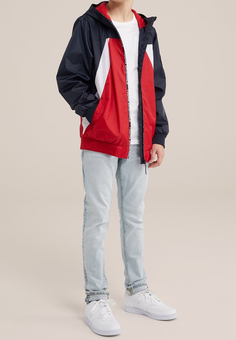 Veste coupe-vent rouge et marine avec des accents blancs, fermeture éclair complète, capuche, poignets et taille élastiques, portée sur une chemise blanche, associée à un jean clair et des baskets blanches.