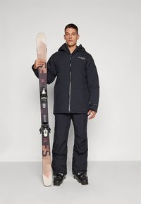 Tumesinine suusajope lukuga, sobitatud suusapükstega. Isik hoiab mustritega suuski, millel on mustad kinnitused ja saapad.