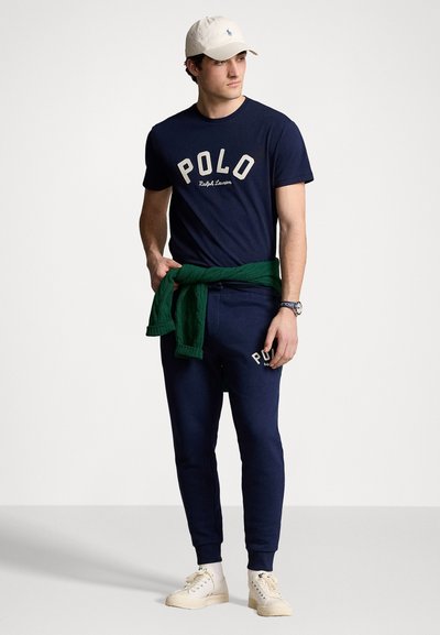 Camiseta de algodón azul marino con el texto "POLO"; pantalones de chándal azul marino a juego; suéter verde atado a la cintura; zapatillas blancas; gorra beige.