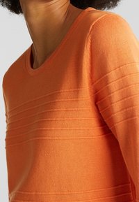 Oranger Strickpullover mit Rundhalsausschnitt und horizontalen Rippdetails über der Brust und an den Ärmeln. Weiche Textur und mittelgroßer Schnitt.