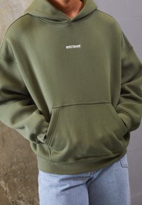 Personne portant un sweat à capuche vert olive avec le texte "WRSTBHVR" sur la poitrine et un jean bleu clair, mains dans la poche avant du sweat.