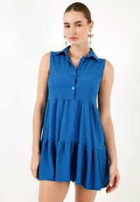 Rochie fără mâneci, de culoare albastră, cu guler cu nasturi, țesătură texturată, margine cu volane stratificate și croială lejeră. Culoare vie și design casual.