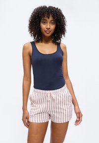 Kvinne med krøllete hår iført en marineblå tanktopp og lyserosa stripete shorts med snor, stående foran en ensfarget hvit bakgrunn.