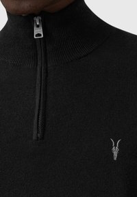 Sort strikket sweater med højt krave og halvlynlås foran. Har en lille grå gedeskalddybde broderi på brystet. Blød tekstur.
