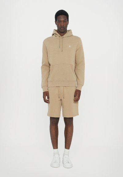 Beige hoodie with front pocket, drawstring hood, and logo; paired with matching beige shorts. Styled with white sneakers.