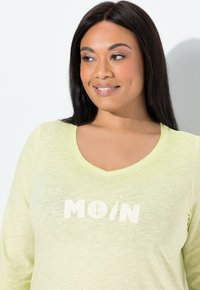 Bluzka z długim rękawem i dekoltem w serek w jasnym zielonym kolorze, wykonana z teksturowanej tkaniny, z białym nadrukiem graficznym zawierającym słowo "MOIN" i motyw kotwicy.
