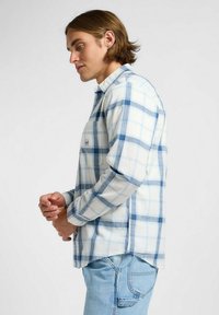 Camisa de franela a cuadros en azul claro y blanco, con mangas largas, cuello abotonado, bolsillo en el pecho izquierdo y aberturas laterales en el dobladillo.