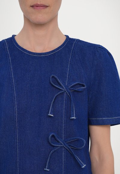 Blue denim top with short sleeves, featuring two decorative bows on the left side and contrasting white stitching details.