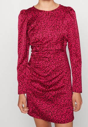 Woman wearing a long-sleeve, fitted, ruched mini dress with a red and dark pink spotted pattern, standing against a plain background.