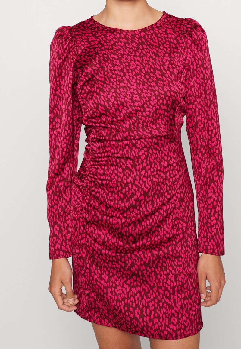 Femme portant une mini-robe cintrée et froncée à manches longues, avec un motif à pois rouge et rose foncé, debout devant un fond uni.