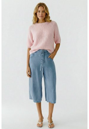 Vrouw draagt een lichtroze, pluizige trui, lichtblauwe, wijde, crop jeans met hoge taille en strikceintuur, en witte sandalen met open teen.